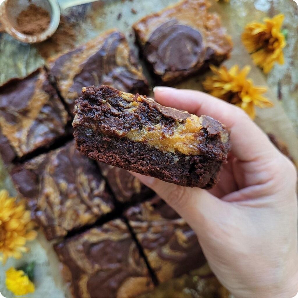 close up view of a slice of caramel brownies. hand is holding the brownie slice so you can see the contrasting layers of brown and amber. background is cut up brownies and yellow fall flowers. 