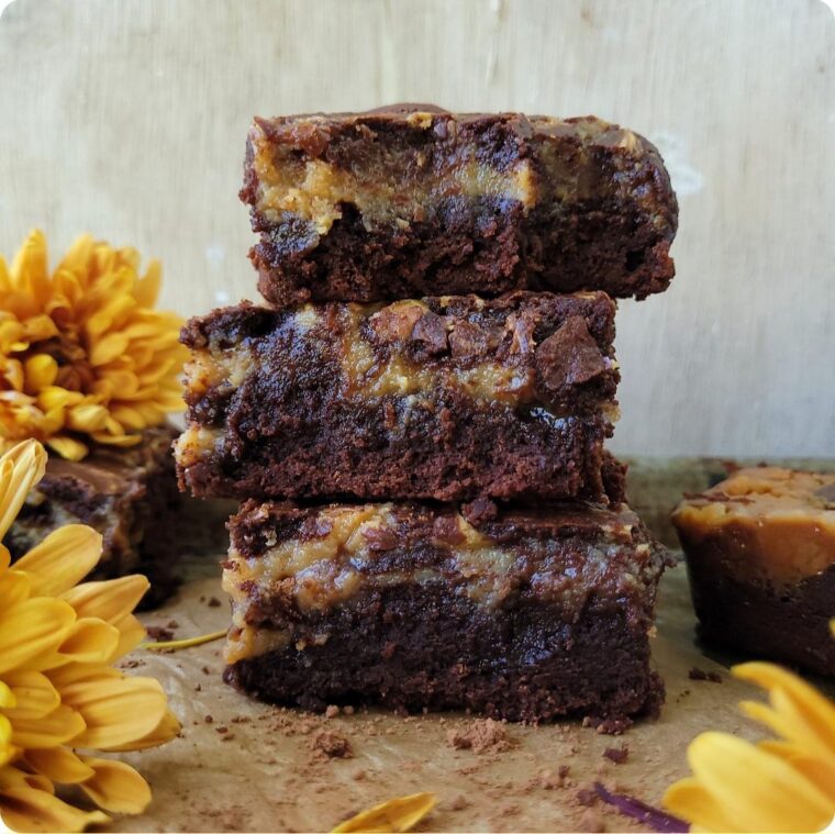 three slices of caramel brownies stacked atop each other. scene is styled with yellow fall flowers. side view image shows gooey caramel layer mixed in with fudgy brownie layer.