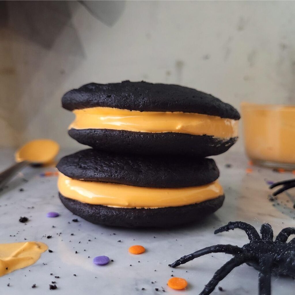 stack of two chocolate halloween whoopie pies. filling is orange marshmallow fluff for contrasting halloween colors. image is styled with spiders and holiday confetti sprinkles. 