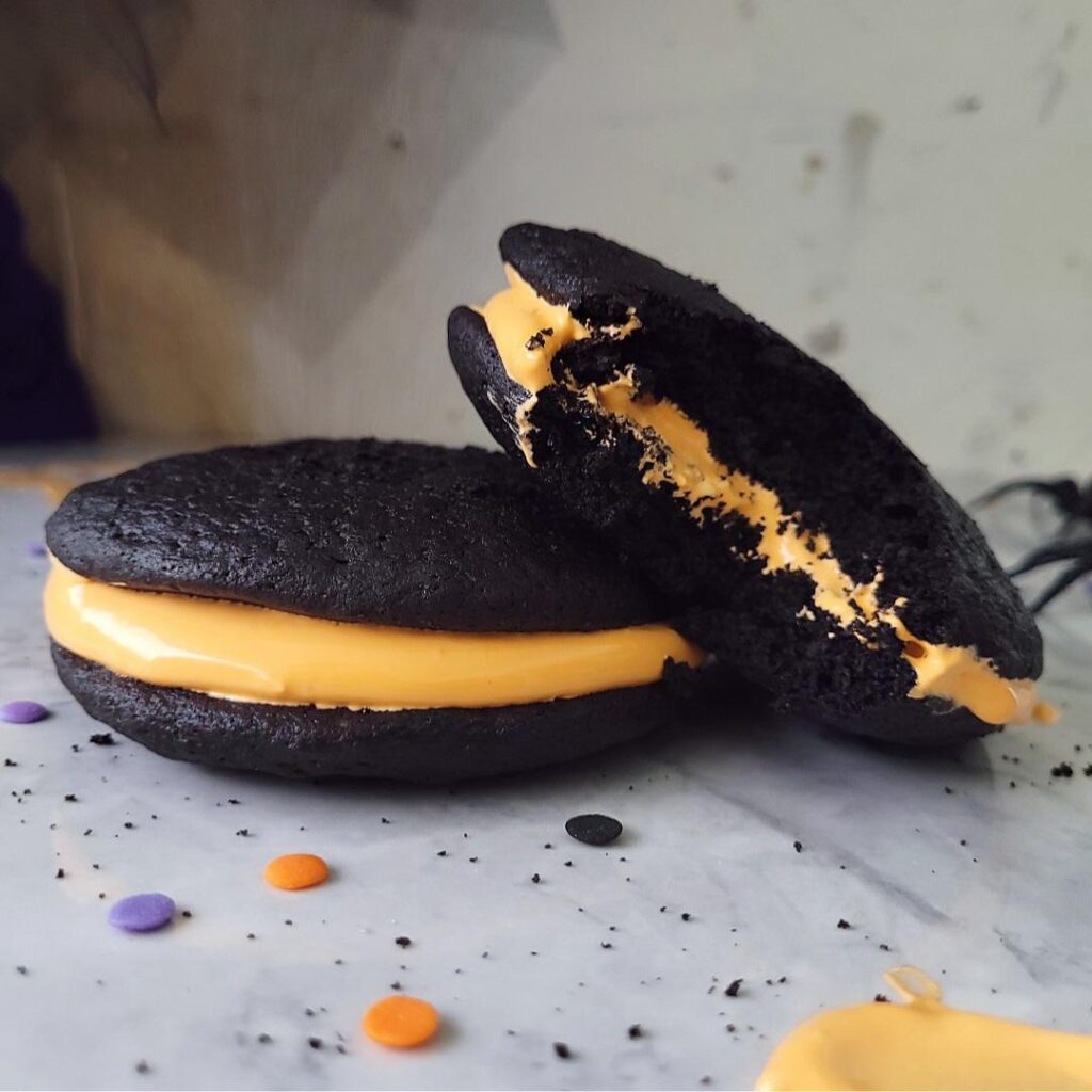 halloween whoopie pies. side view of one dark chocolate whoopie pie with orange marshmallow filling leaning on another pie. the leaning pie has been bitten so you can see the soft inner crumb. 