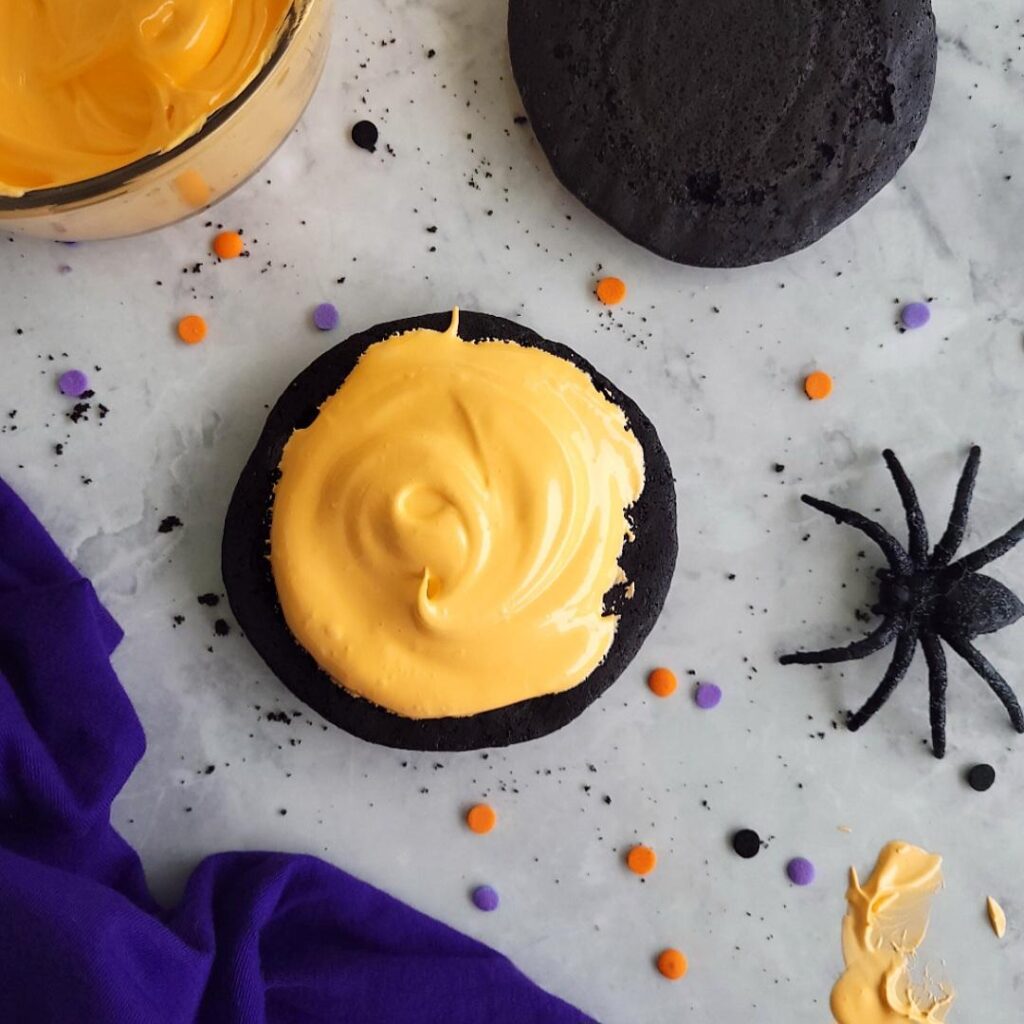 halloween whoopie pies. top down shot of whoopie pie in the assembly phase. orange marshmallow filling has been added but not the top of the cookie. shot is styled with halloween confetti sprinkles a black spider and purple linen. 