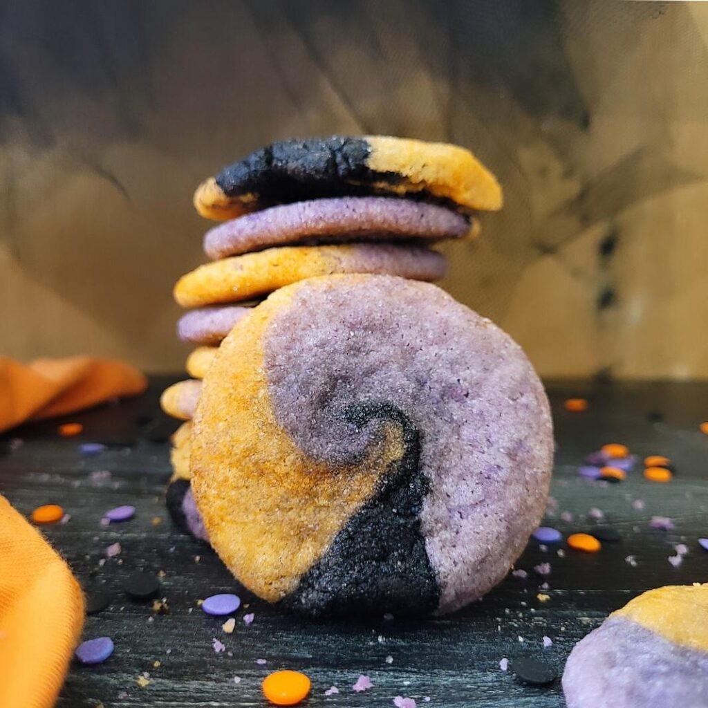 hocus pocus cookies. stack of halloween sugar cookies colored orange purple and black. one cookie is standing straight up and the black swirl looks like a witch hat. 