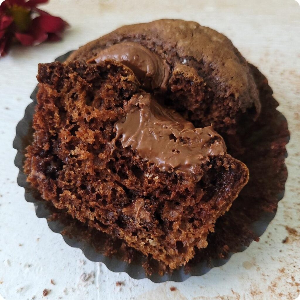 chocolate nutella muffins. close of view of one muffin that has been cut in half so you can see the moist inner crumb. top of muffin has a swirl of nutella. muffin is in a black cupcake liner. 