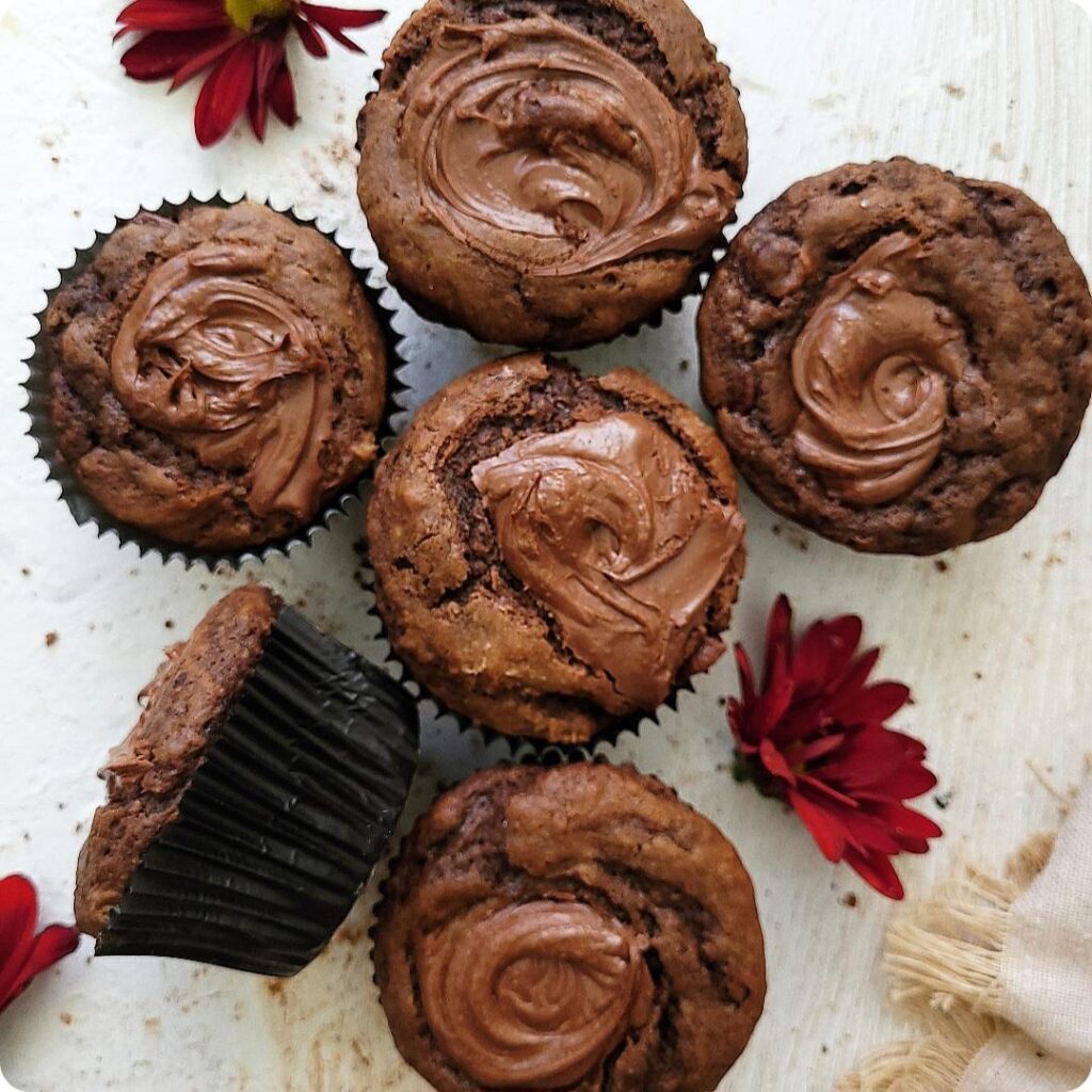 nutella muffins. top down view of muffins with a swirl of nutella on top. background is white wood and image is styled with maroon fall flowers.