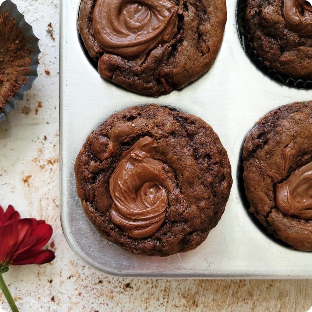 chocolate nutella muffins top down view of baked muffins in a muffin tin. muffins have a swirl of nutella on top. 