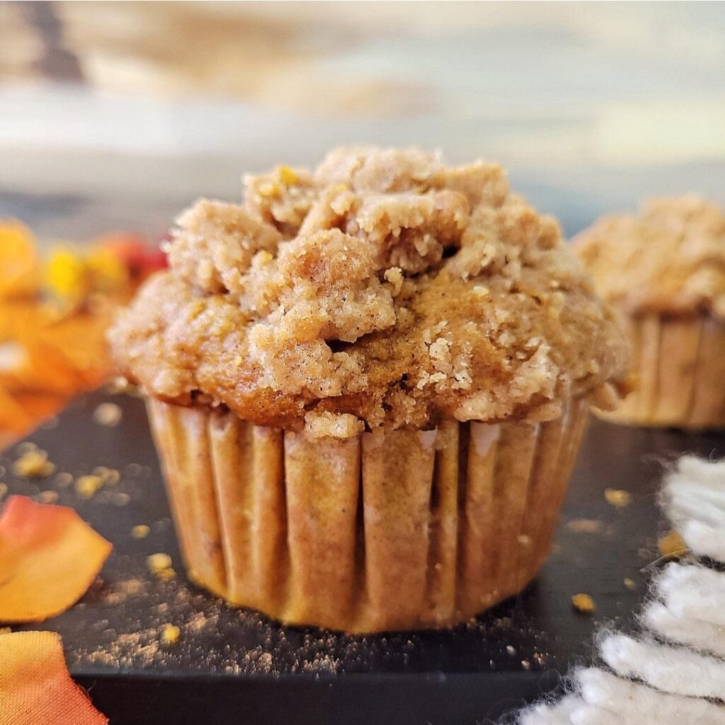 pumpkin banana muffins. close up view of a single muffin on black wood. frame is styled with fall colored leaves. muffin has a crumb topping and a brown muffin liner. 