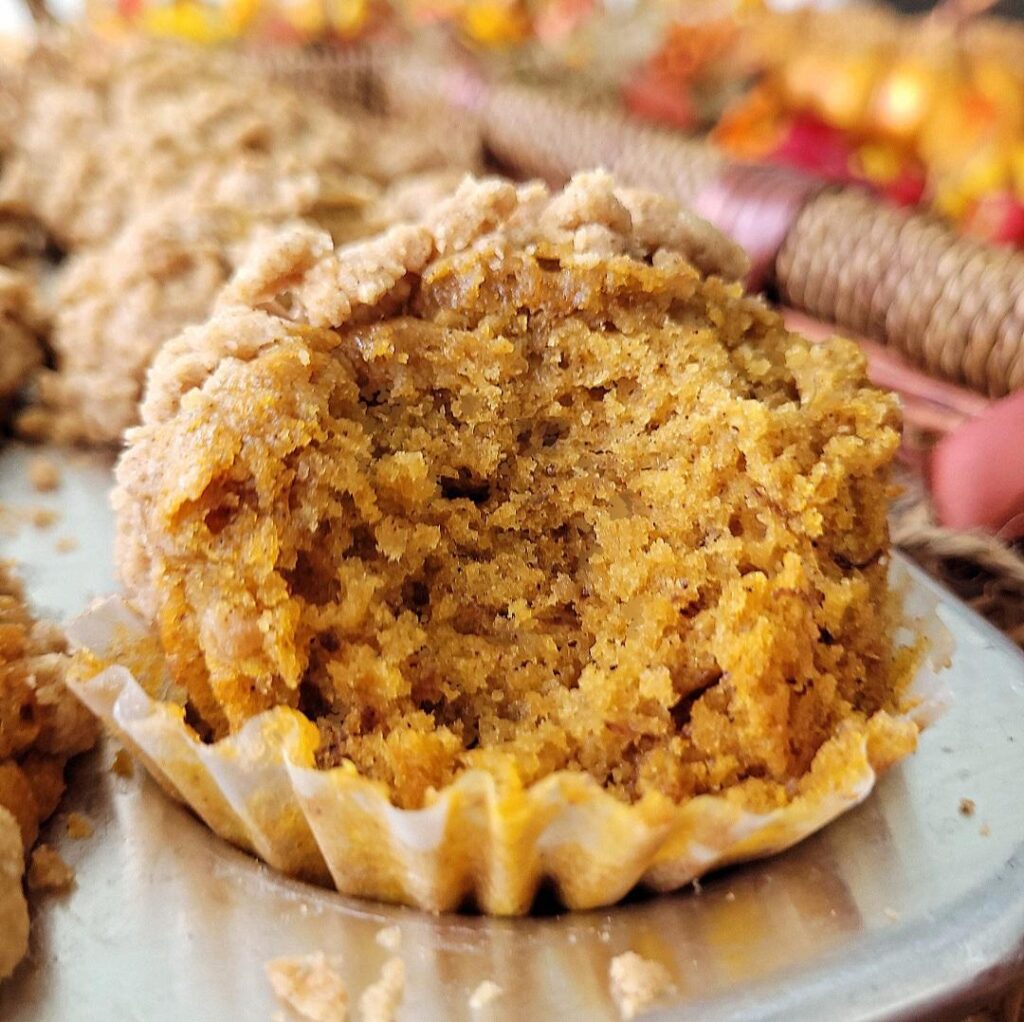pumpkin banana muffin with crumble topping in a muffin tin. muffin has been bitten so you can see the inner pumpkin colored crumb.