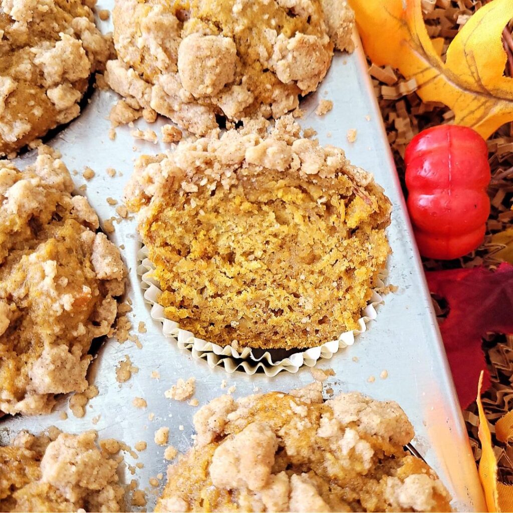 pumpkin banana muffins with crumb topping. muffins are in a muffin tin. one muffin has been cut in half so you can see the pumpkin colored crumb. frame is styled with fall colored leaves.