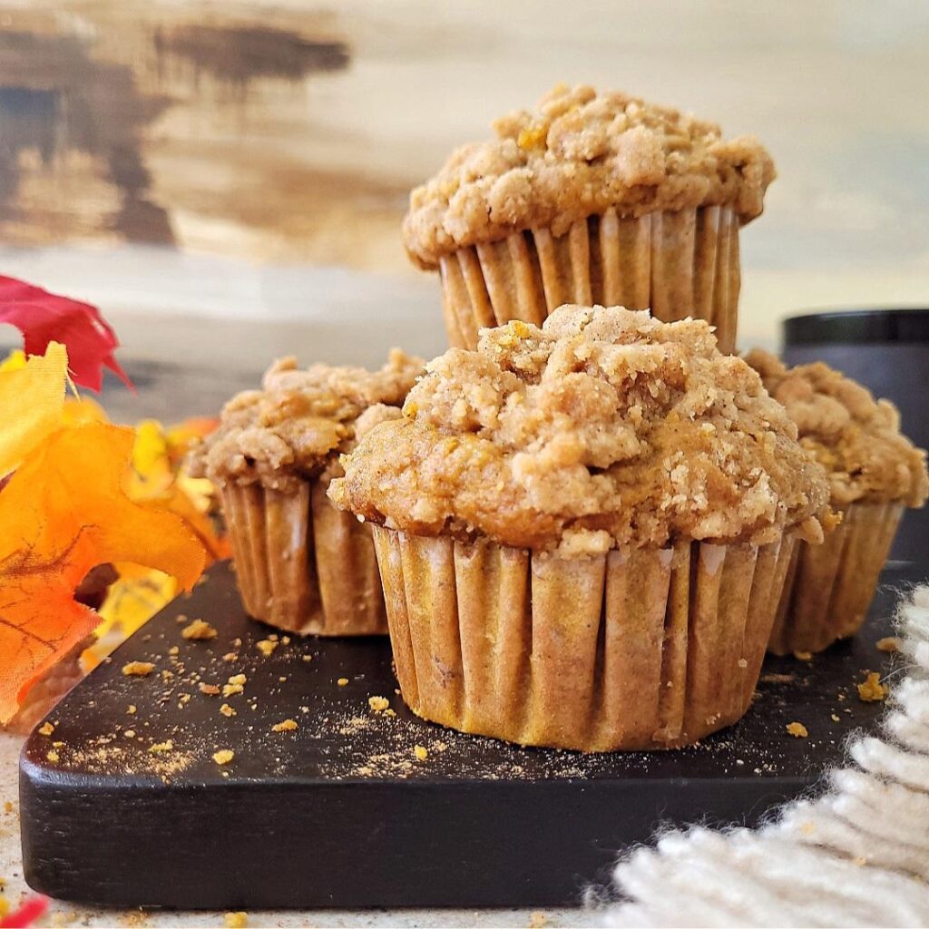 pumpkin banana muffins with crumb topping. side view of muffins on a black wood. 4 muffins are visible, three in a V pattern and one stacked high in the back. frame is styled with fall colored leaves. 