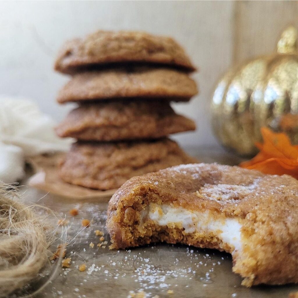 pumpkin cheesecake cookies. side view. cookie in front of frame is bitten so you can see the cheesecake center. back of frame is a 4 stack of cookies and a golden pumpkin decoration. 