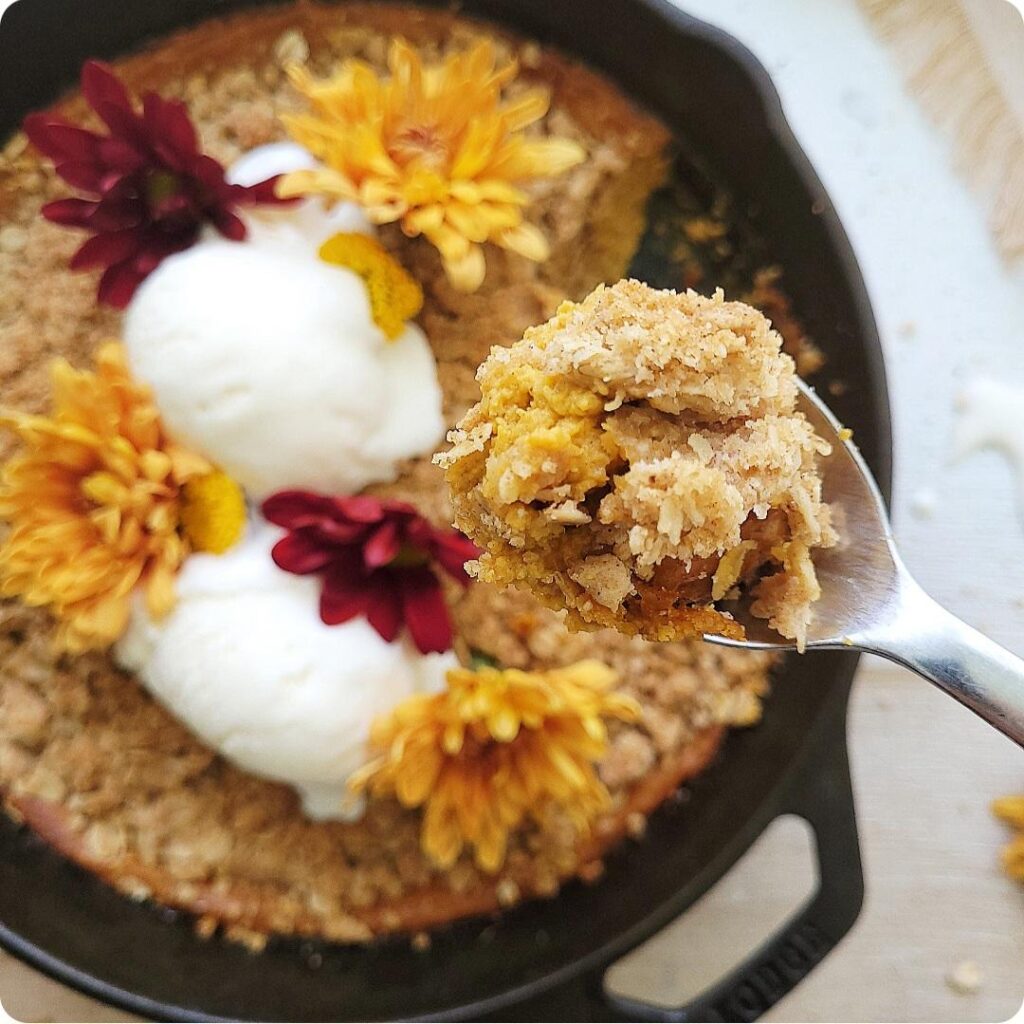 pumpkin crisp baked in a cast iron skillet. top down view of crisp garnished with scoops of vanilla ice cream and fall flowers. skillet is in the background out of focus, a spoonful of the pumpkin crisp is zoomed in center frame. 