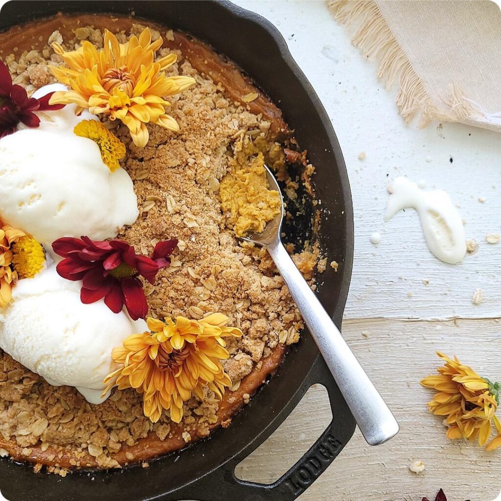 pumpkin crisp baked in a cast iron skillet. top down view of crisp garnished with two scoops of vanilla ice cream and fall flowers. part of the crisp has been scooped out and there is a dirty spoon in the void. 