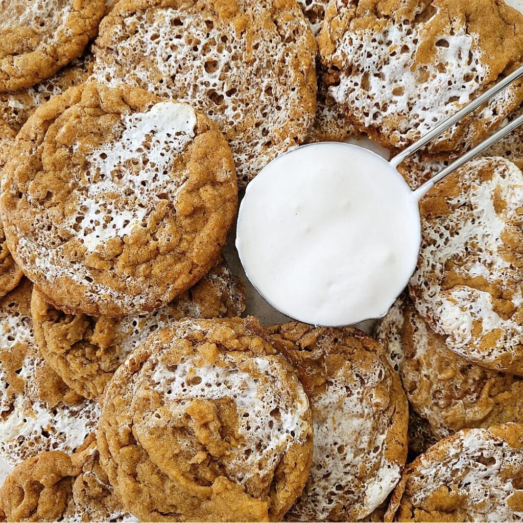 pumpkin marshmallow cookies. top down view of a pile of cookies surrounding a measuring cup filled with marshmallow creme. 