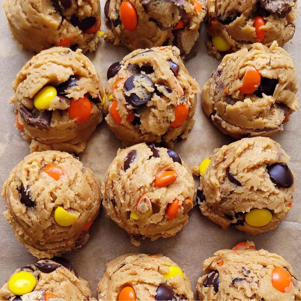 close up view of reese's pieces cookie dough balls on a brown piece of parchment paper. dough balls are tightly packed and touch each other. dough is loaded with dark chocolate chips, colorful reese's pieces candies and peanut butter cup pieces. 