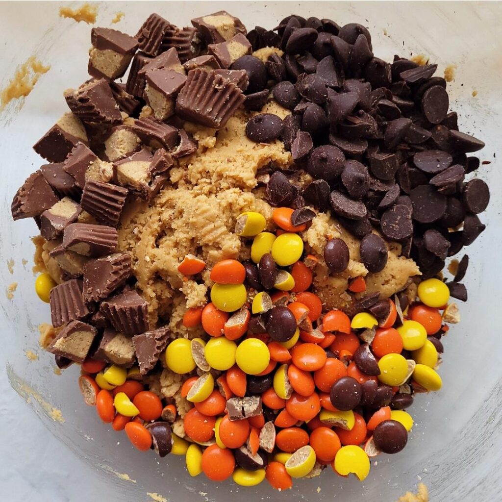 reese's pieces cookies. top down view of unmixed dough with piles of reese's pieces, chopped reese's peanut butter cups and dark chocolate chips. 