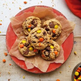 reese's pieces cookies. five cookies on brown parchment paper on a red plate. background is cookie crumbs and broken reese's pieces.