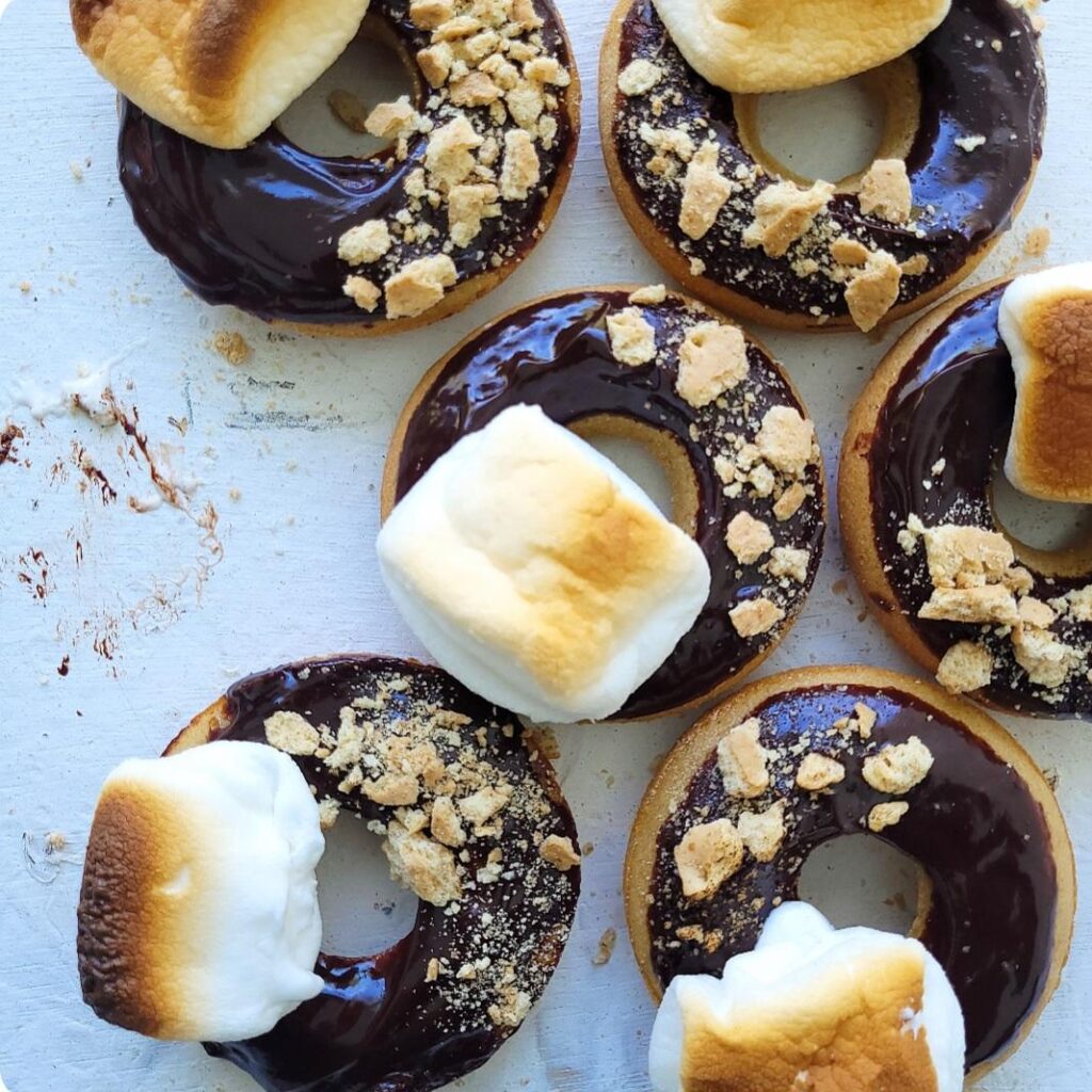 s'mores donuts. top down view of baked donuts topped with chocolate ganache, toasted marshmallows and graham cracker crumbs. 