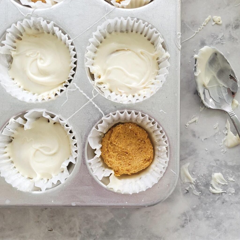 white chocolate pumpkin cups in a silver muffin pan. all cups but one have been coated with white chocolate and there is a chocolate covered spoon next to the pan. 