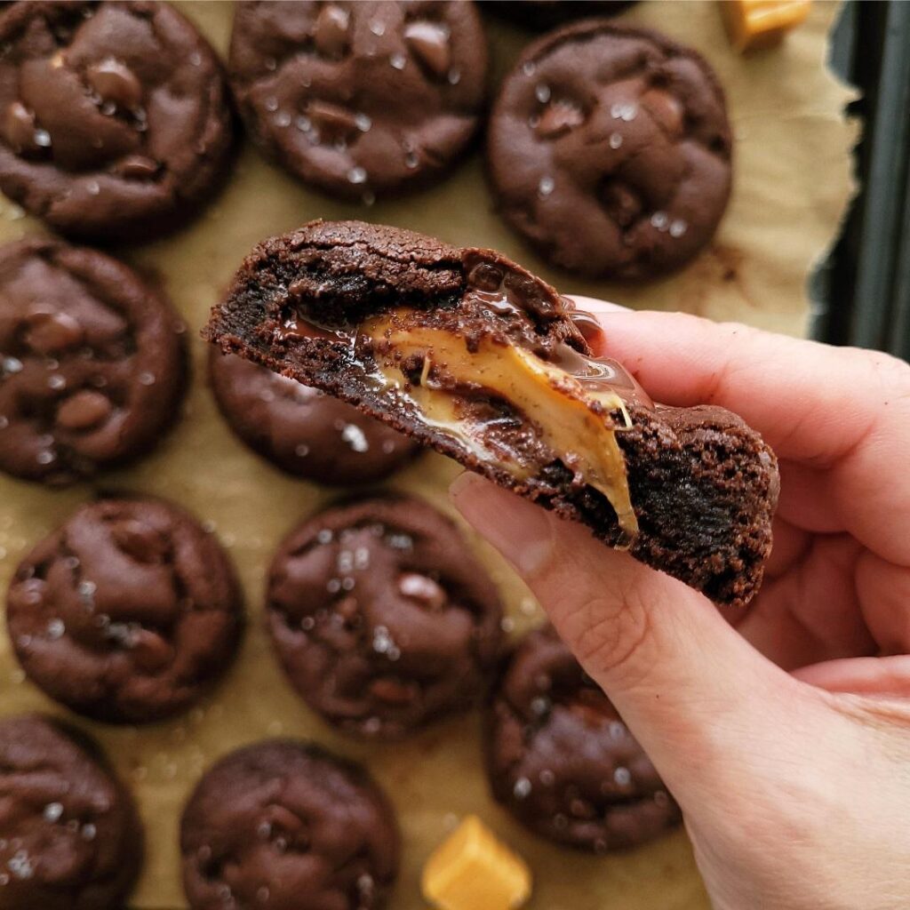 chocolate caramel cookies. close up view of a hand holding a caramel stuffed chocolate cookie. cookie is cut in half so you can see the gooey caramel center. background is a baking tray filled with cookies. 