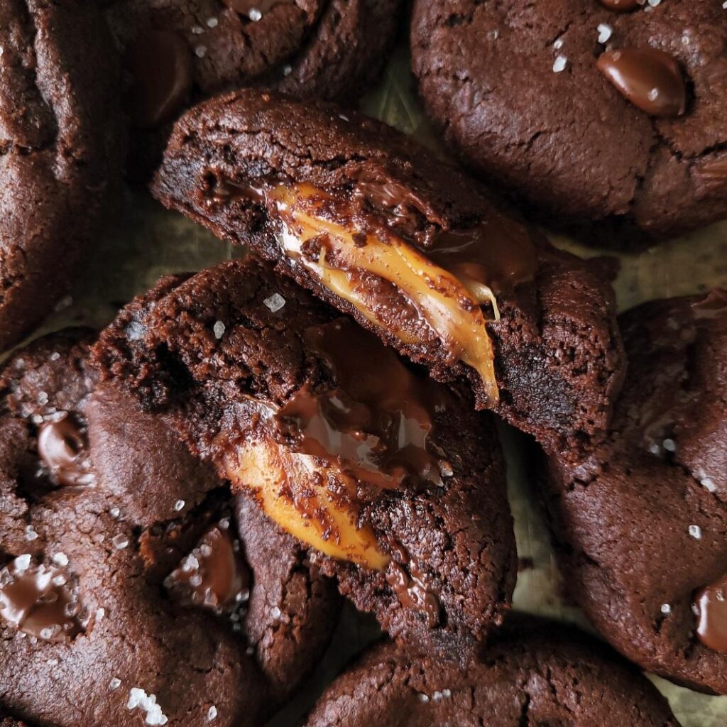 chocolate caramel cookies. top down view of dark chocolate cookies stuffed with caramel. one cookie is cut in half and facing upwards so you can see the gooey caramel center. these cookies are surrounded by more chocolate cookies that are topped with chocolate chips and sea salt.