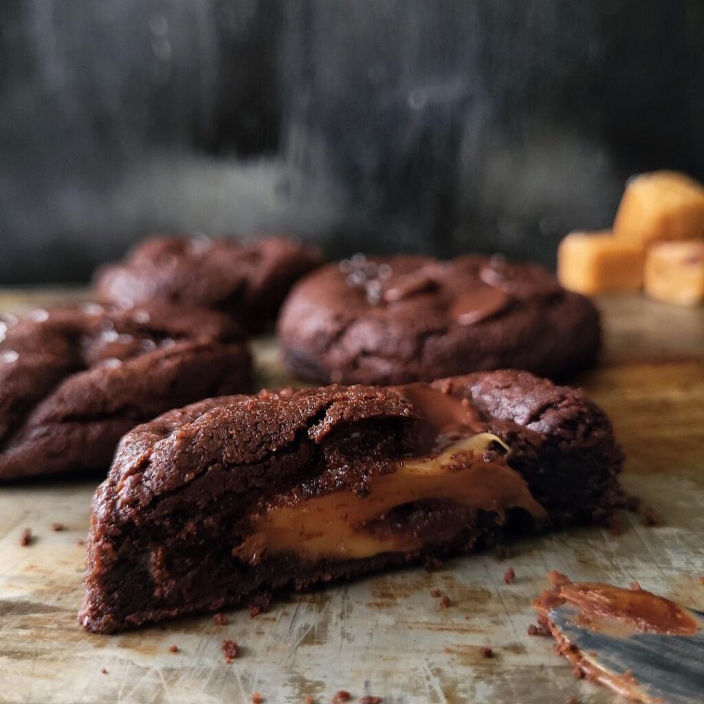 chocolate caramel cookies. side view of four cookies on a distressed tin surface. the cookie in front is cut in half so you can see the gooey caramel center. 