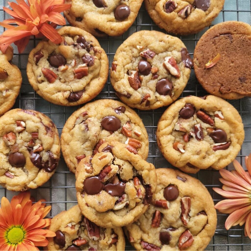 chocolate chip pecan cookies. top down view of cookies scattered on a baking rack. cookies are topped with melty chocolate chips and chopped pecans. frame is styled with orange fall flowers. 