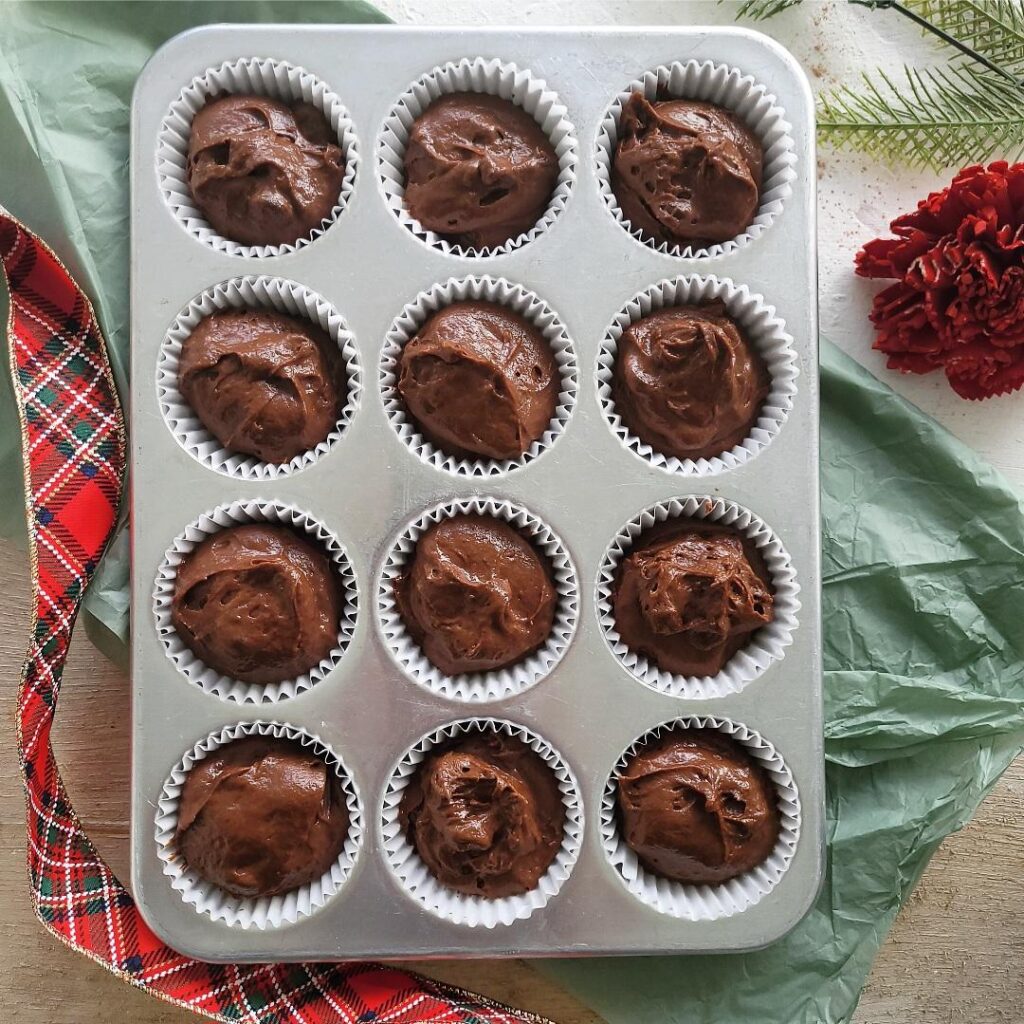 chocolate gingerbread muffins. top down view of unbaked batter in a silver muffin pan. scene is styled with red christmas ribbon, a red flower and greenery from a christmas tree. 