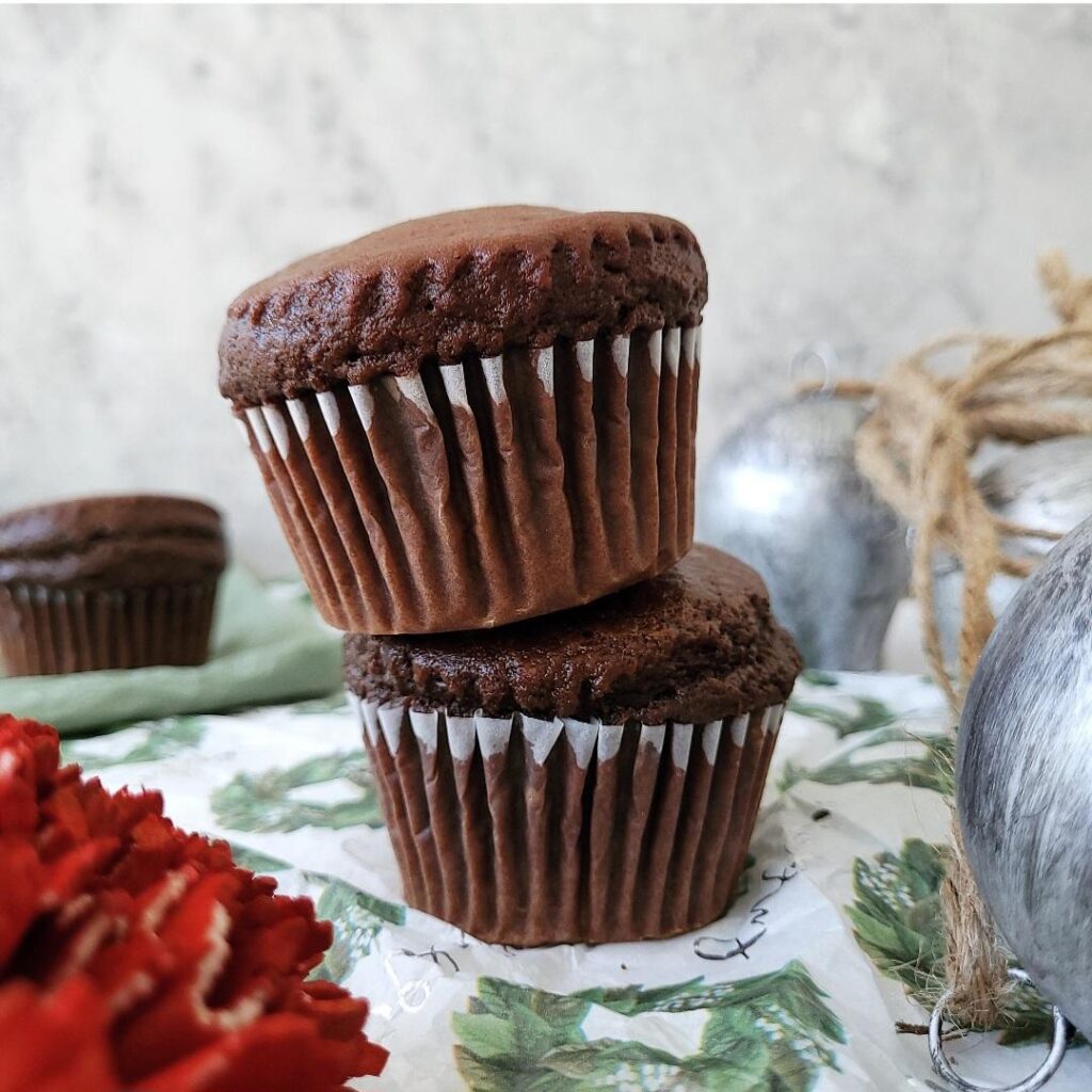 chocolate gingerbread muffins. side view of two muffins stacked on top of each other. image is styled with silver christmas bells. 