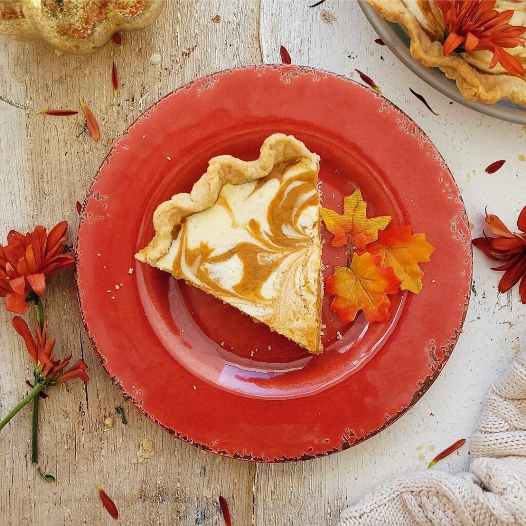 cream cheese pumpkin pie. top down view of a slice of pumpkin pie swirled with cream cheese on a red plate with tiny fall leaves. 