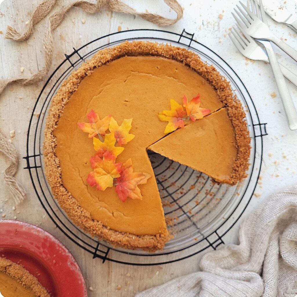 graham cracker crust pumpkin pie. top down view of pie with one slice missing and another slice cut. top of pie is styled with tiny fall leaves. the glass pie dish is sitting on a round wire cooling rack that is black. 