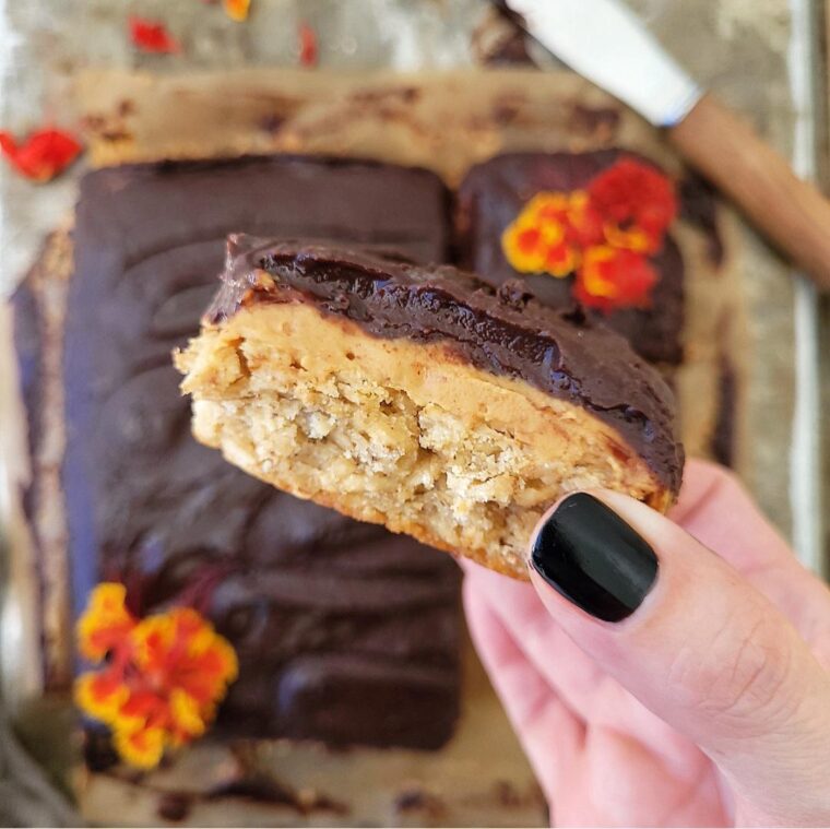 chocolate peanut butter oatmeal bars. close up view of a hand holding a cut bar so you can see the oatmeal, peanut butter and chocolate layers. the rest of the batch can be seen in the blurred background