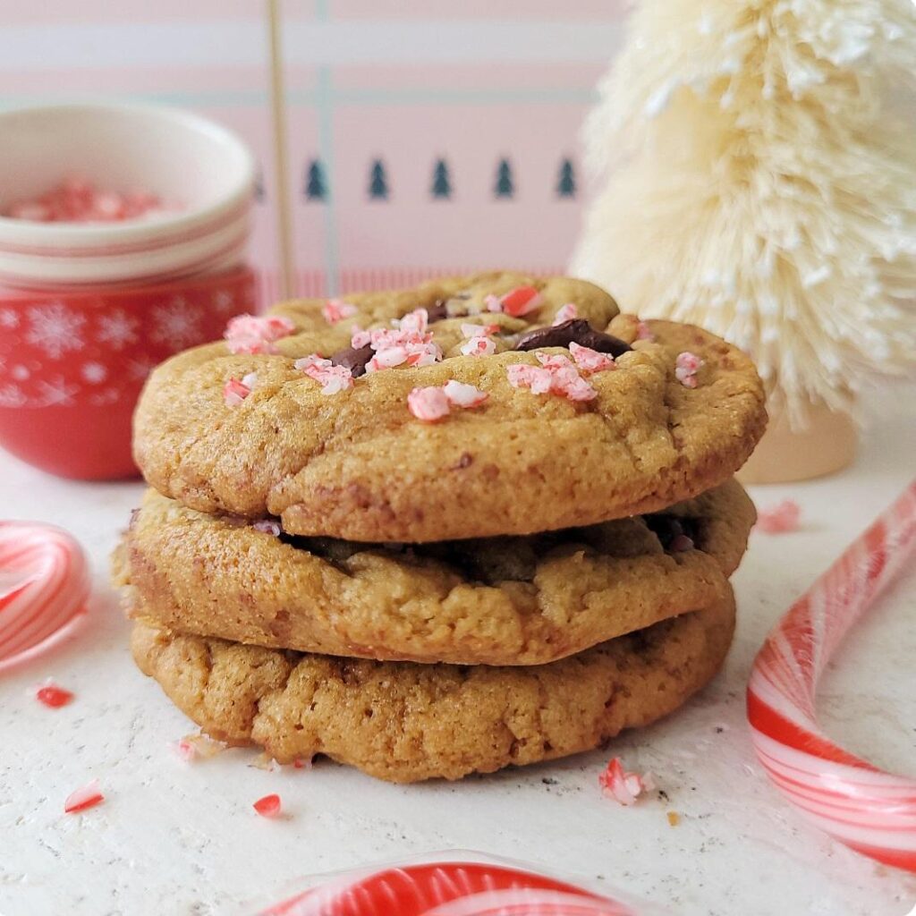 peppermint chocolate chip cookies. side view of three cookies stacked on top of each other. top cookie is topped with broken candy canes and chocolate chips. background is pink chrismas paper, a white bottlebrush tree and small red bowls with snowflakes. 