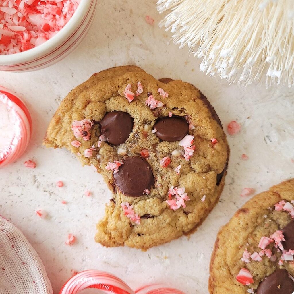 peppermint chocolate chip cookies. top down view of a cookie topped with three large chocolate chips and bits of broken candy cane. a bite has been eaten from the cookie.
