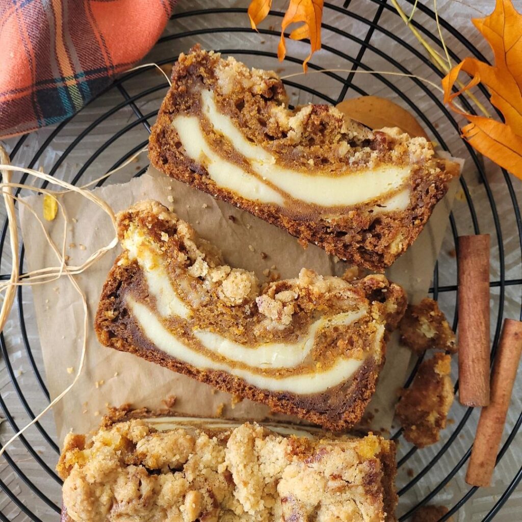 pumpkin cream cheese bread. top down view of loaf on a round black baking rack. two slices have been cut and are laying flat so you can see the inner swirls of cream cheese with pumpkin bread