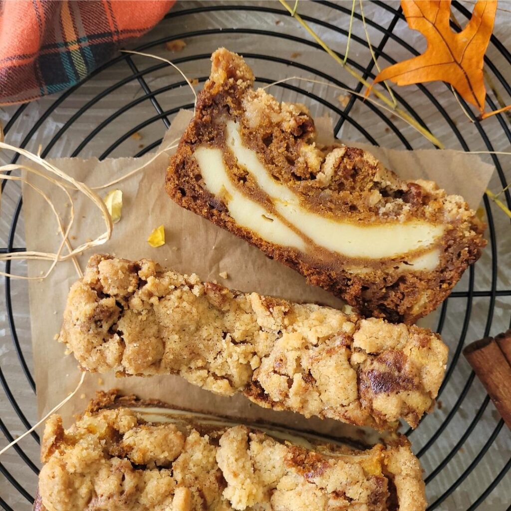 top down view of a loaf of pumpkin cream cheese bread on a round baking rack. bread has two slices cut from loaf. one slice is upright and the other is on it's side so you can see the inner cream cheese swirl. 