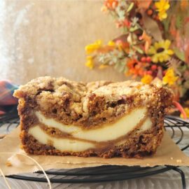 pumpkin cream cheese bread with cinnamon crumb topping. side view of a cut loaf so you can see the inner pumpkin crumb swirled with two layers of cream cheese. background is distressed wood and colorful fall flowers.