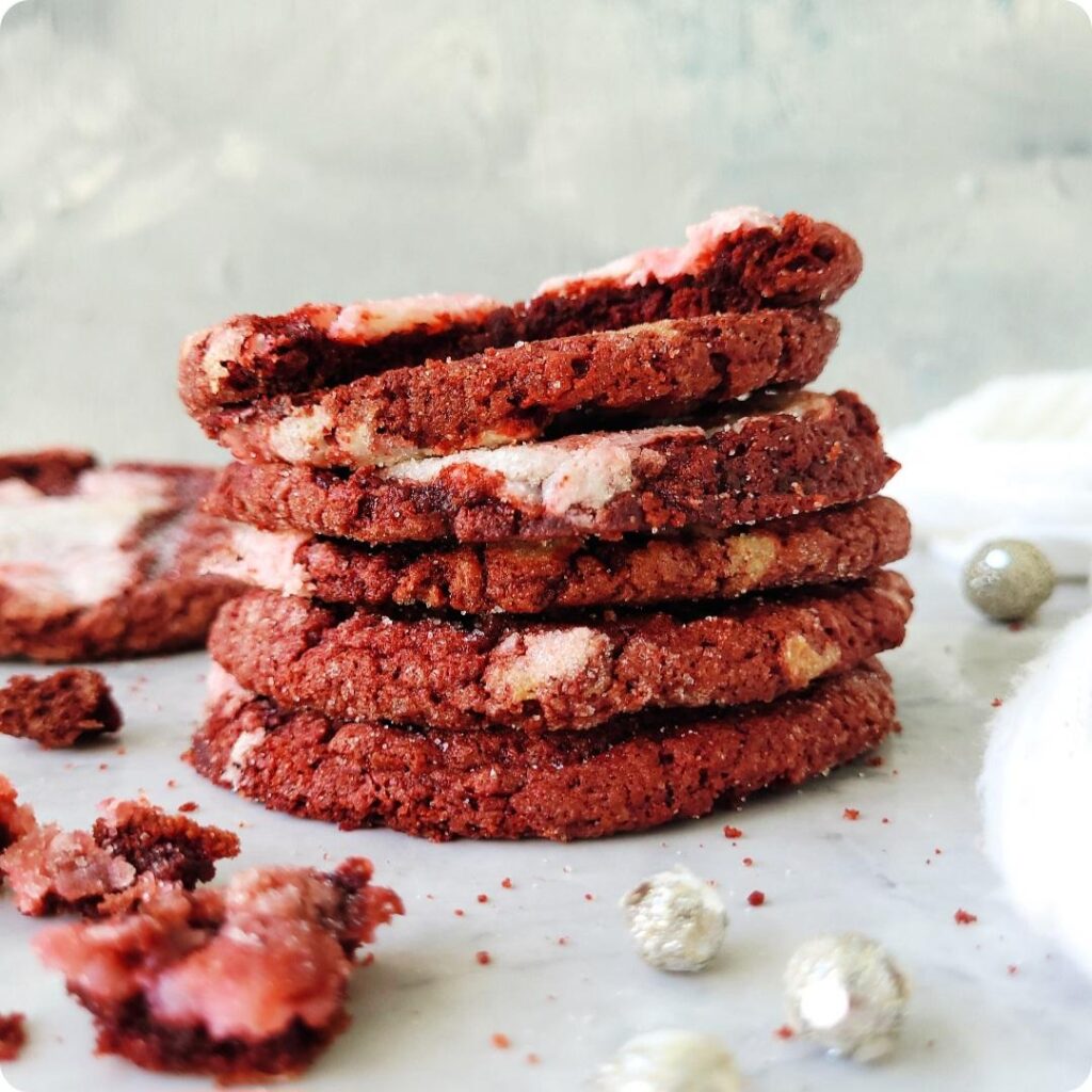 red velvet cookies. side view photo of a stack of 6 cookies. top cookie is bitten so you can see the inner red crumbs. surface and background is gray marble. 
