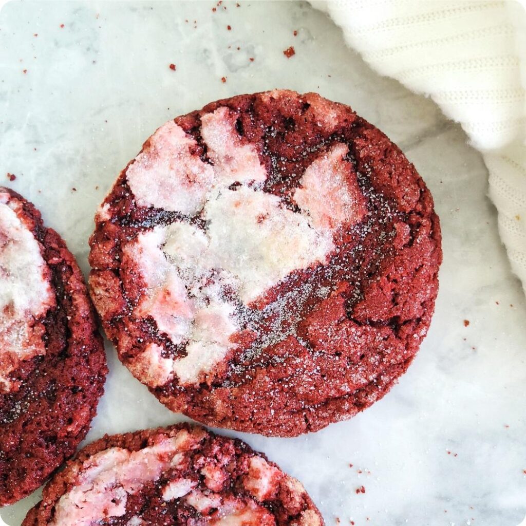crackly red velvet cookies with cream cheese baked in. top down view of a single centered cookie on a marble surface. two partial cookies are visible in the lower left corner.