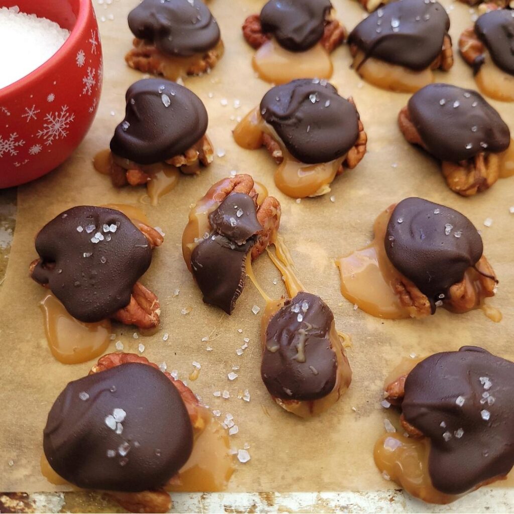 caramel pecan turtles. top down view of candy turtles on brown parchment paper. turtles have been sprinkled with sea salt. one turtle is broken in half and has gooey caramel between the pieces. 