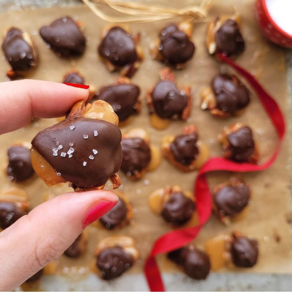 chocolate pecan turtles. close up view of aa hand holding a turtle that's been sprinkled with sea salt. background is a cookie sheet filled with turtles on brown parchment paper. image is styled with red ribbon and a red bowl of sea salt. 