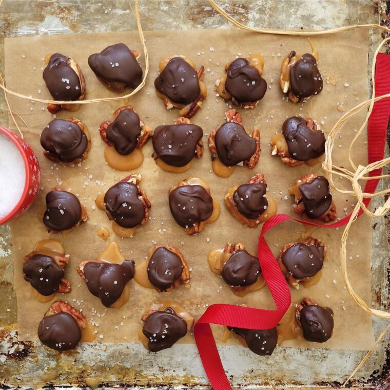 homemade pecan turtles. top down view of turtle candy lined up in rows on brown parchment. styled with a red ribbon and a small red bowl of sea salt.