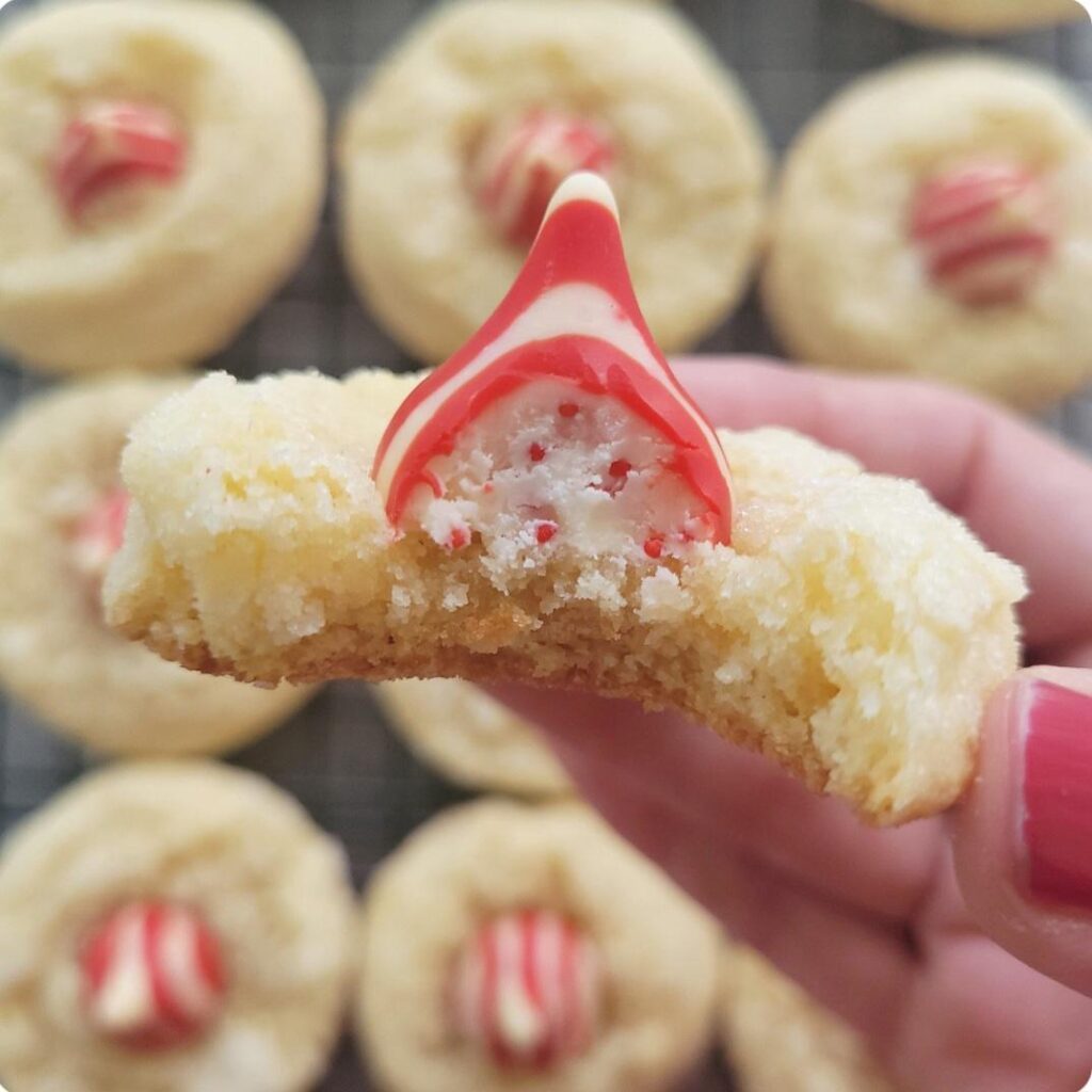 peppermint kiss cookies. close up view of a hand holding a peppermint kiss sugar cookie with a bite taken out of it so you can see the inner crumb and the inner red and white speckled candy cane flavored hershy's kiss. background is blurred cookies on a baking rack. 