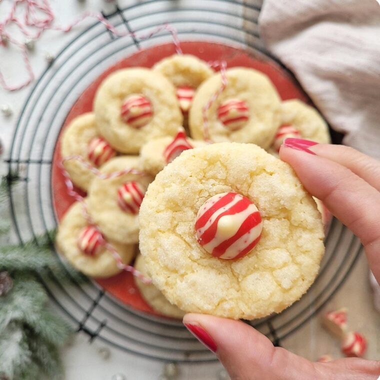 peppermint kiss cookies. top down view of these christmas cookies piled on a red plate in the background. foreground is a hand holding one of the sugar cookies. they are all topped with red and white striped candy cane hershey kisses.