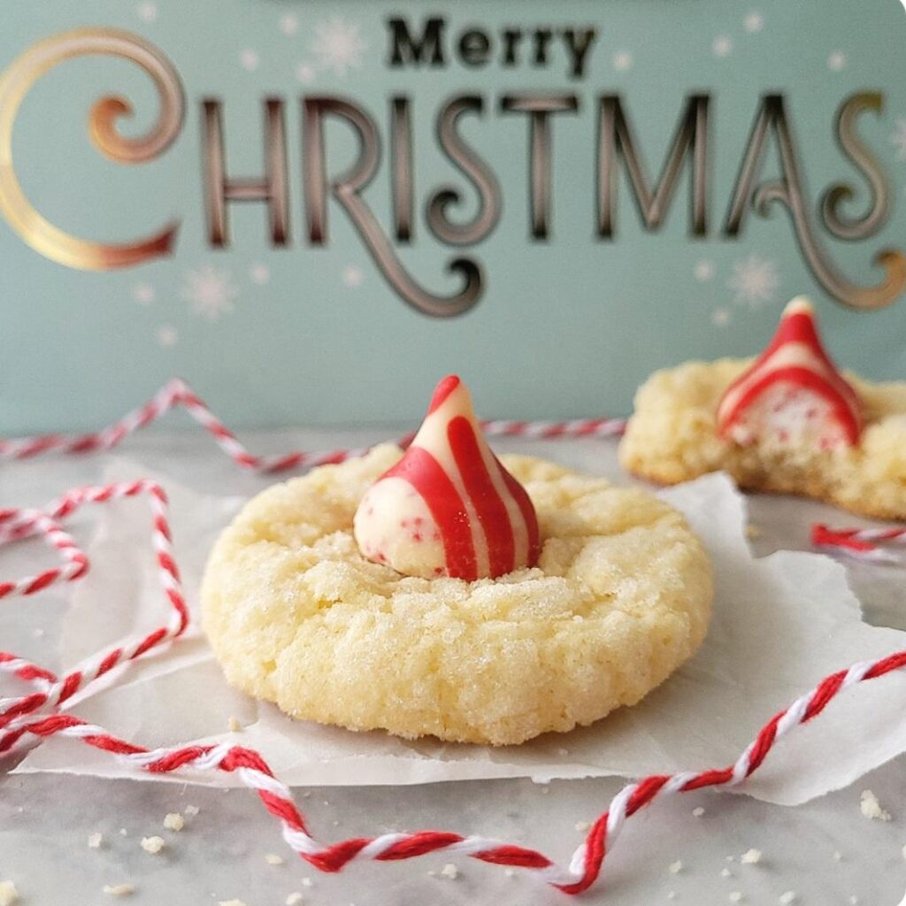 peppermint kiss cookies. side view of two sugar cookies with red and white striped Hershey's kisses on top. background is blue and says merry christmas. 