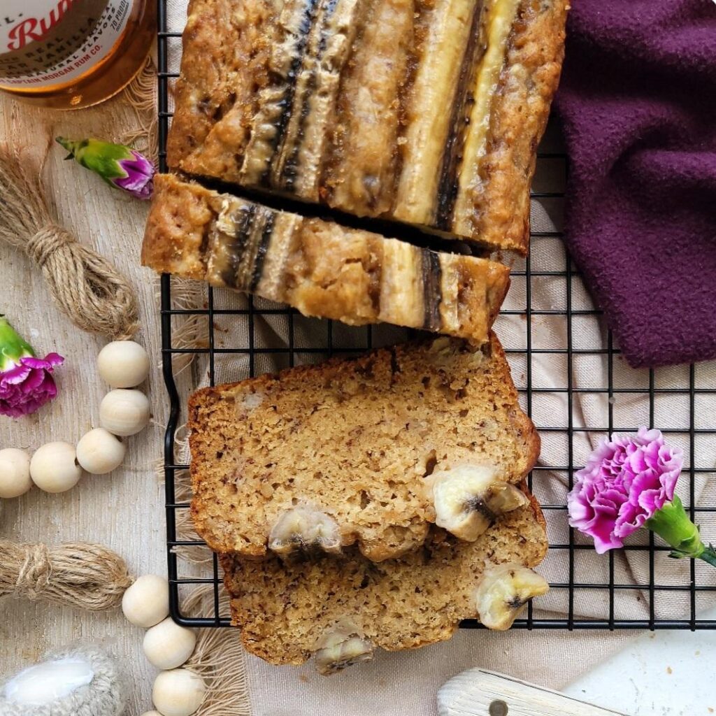 brown butter rum banana bread. top down view of loaf on a black backing rack. two slices are cut from the loaf and lying down. one slice is cut and still standing. image is styled with purple flowers and a bottle of captain morgan's spiced rum. 