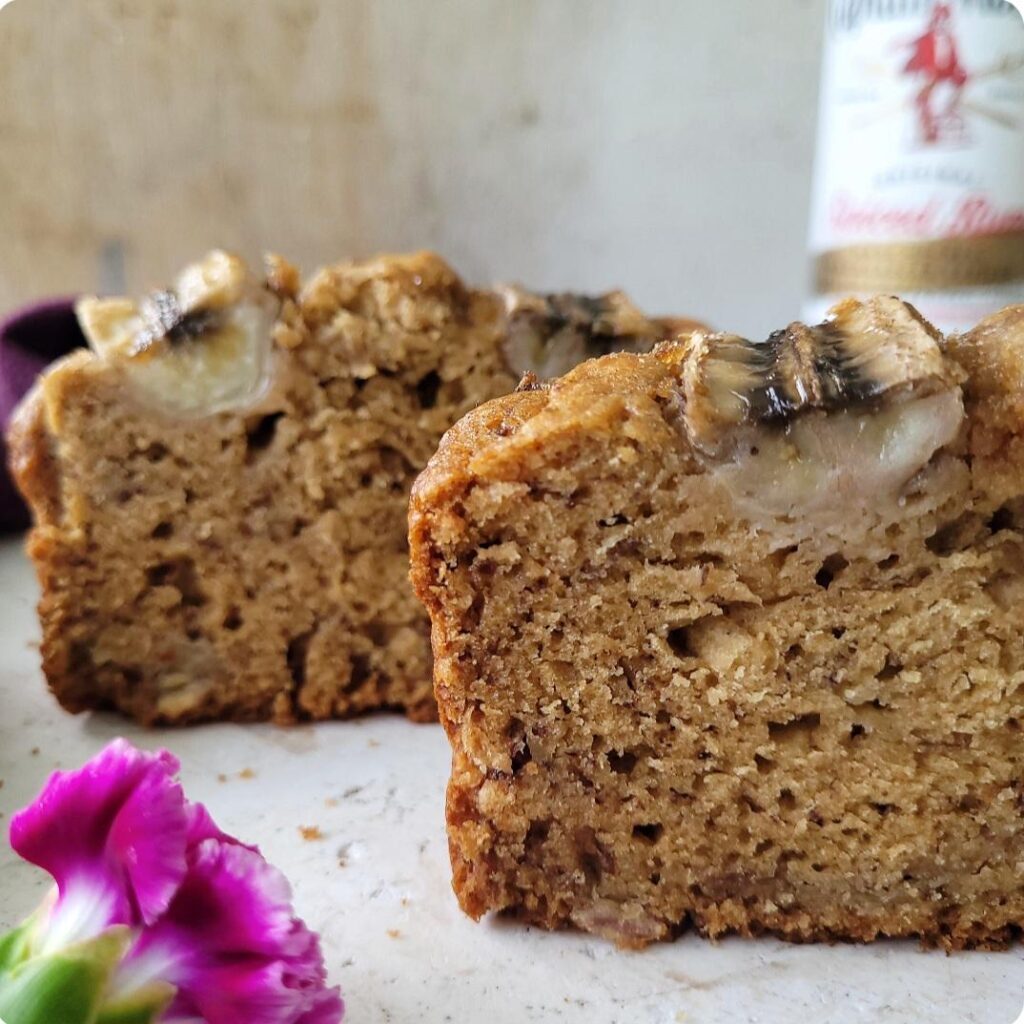 brown butter rum banana bread. side view of two cut slices standing on a white marble surface with a bottle of rum in the background and a purple flower in the foreground. 