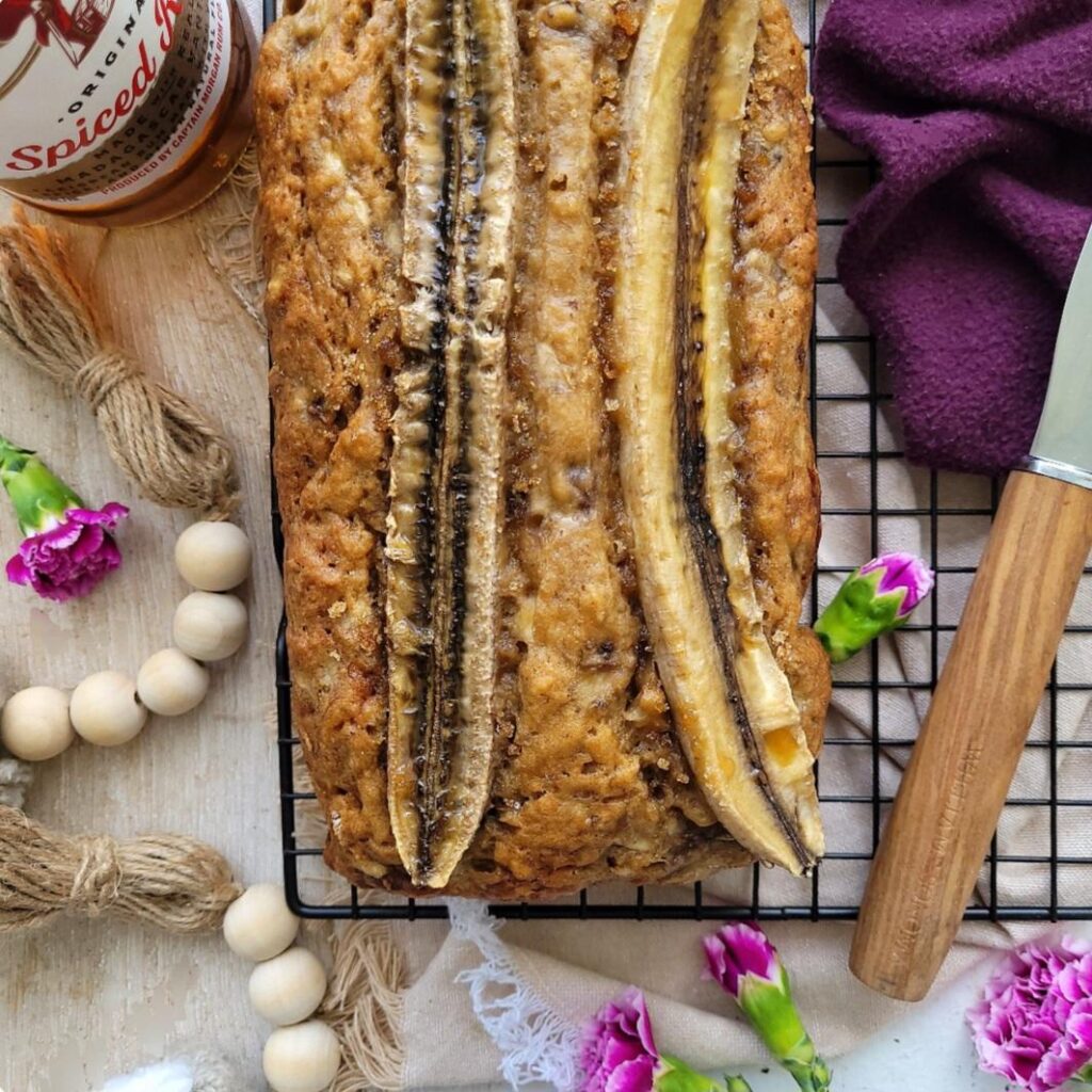 rum banana bread. top down view of an uncut loaf with baked slices of banana on top. image is styled with a bottle of captain morgan's rum, purple flowers and wooden beads.