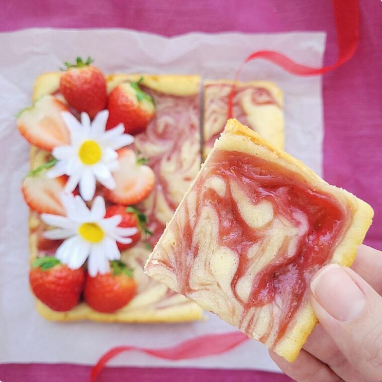 strawberry blondies with swirls of homemade strawberry sauce. top down view of a cut square of blondies close to the camera lens. background is the remaining pan of blondies topped with fresh strawberries and white daisies