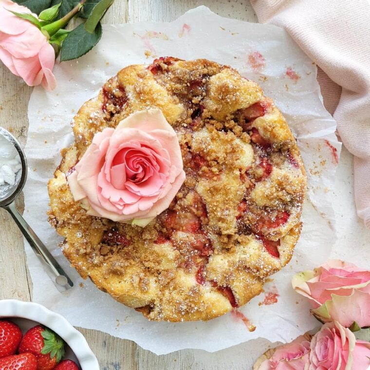 strawberry crumb cake. top down view of baked cake garnished with a large pale pink rose. image is styled with stemmed roses and a bowl of strawberries.