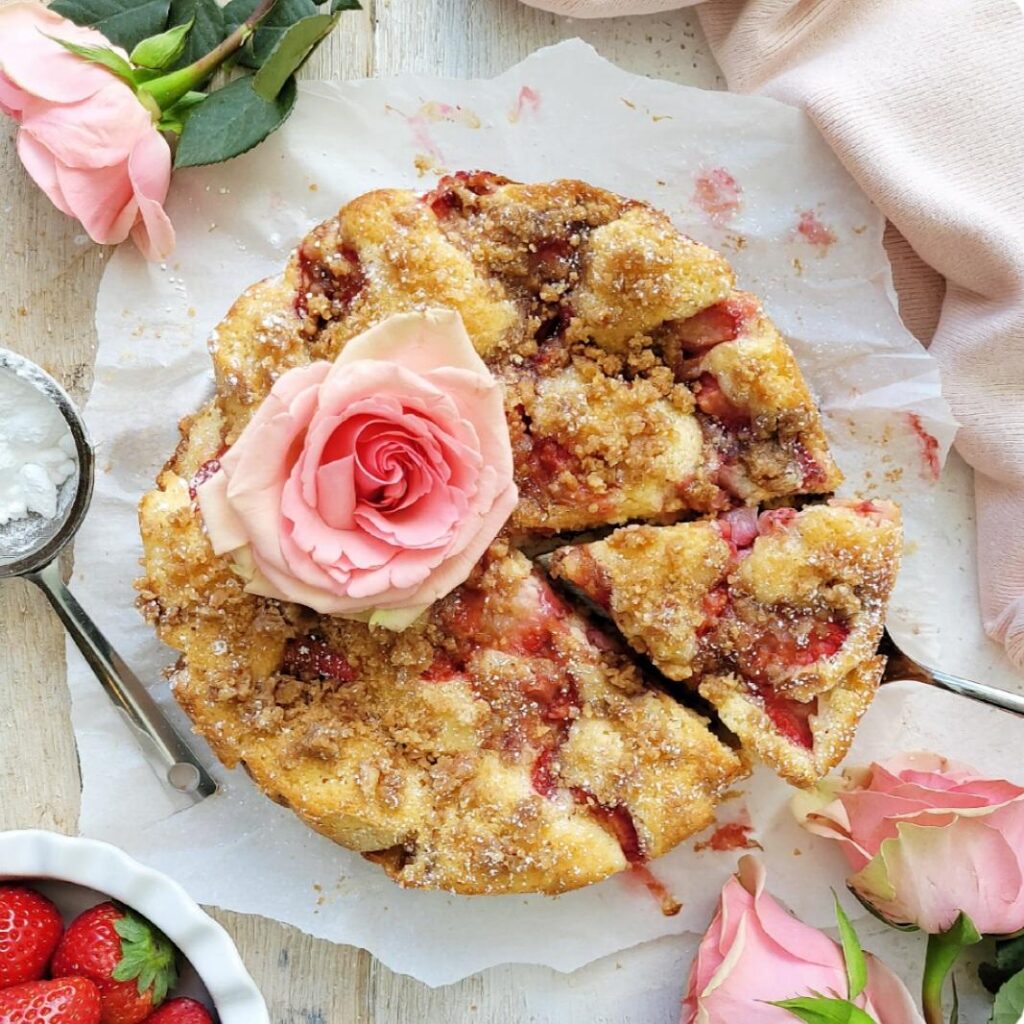 strawberry crumb cake. top down view of cake with a large pale pink rose on top. one slice has been cut and is being removed with a silver cake server.